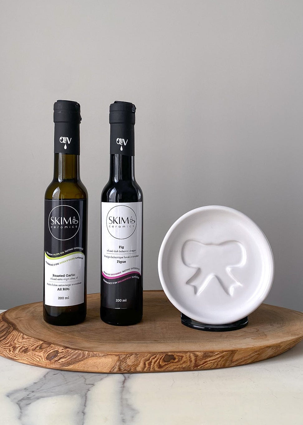 Two bottles of olive oil on a wooden board with a white dish featuring a logo.